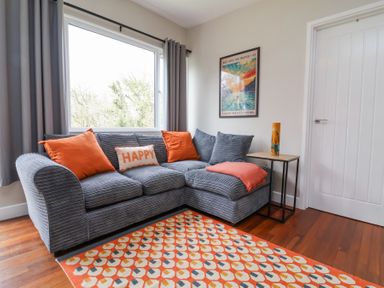 A living room with a grey sectional sofa with orange pillows and a geometric rug at 15 The Park in Kilkhampton