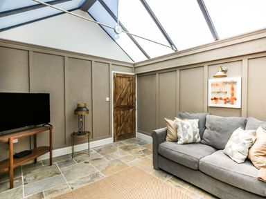 A room with a gray sofa with pillows a wooden door a flat screen tv on a wooden stand and a lamp on a small round table at The Apple Loft at Jordan House in Preston