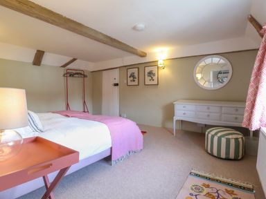 A bedroom with a bed a bedside table with lamp a white dresser a round mirror and a striped pouf at Brook Cottage in Peasenhall