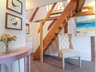 An interior space with wooden stairs beige upholstered chair framed bird prints on the wall and a pale pink table with flowers at Brook Cottage in Peasenhall