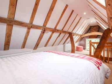 An attic bedroom with exposed wooden beams a white bedspread a pink blanket a green chest of drawers and a pink ottoman at Brook Cottage in Peasenhall