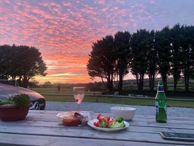 A wooden table outdoors with snacks a glass a bottle and a phone with trees and a sunset sky in the background at Wheal Amelia Lodge in Wendron near Helston