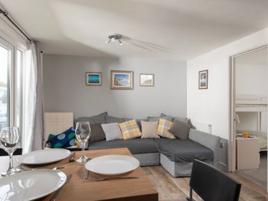 A living room with a grey sectional sofa, cushions, framed pictures on the wall and a dining table with plates and glasses at Wheal Amelia Lodge in Wendron near Helston