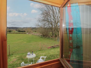A view of a green field with trees and hills seen through an open wooden window at Harri's Hut in Bishop's Lydeard