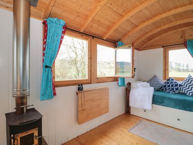 A wooden room with a stove a foldable wall table a day bed with cushions and windows with blue curtains at Harri's Hut in Bishop's Lydeard