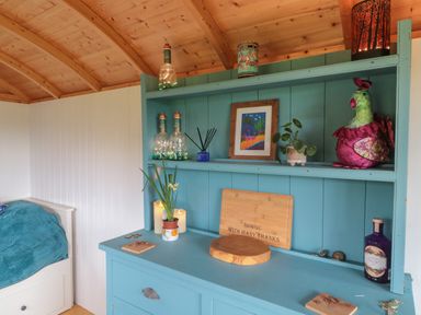 A blue wooden shelf with bottles plants framed picture and decorative items next to a bed at Harris Hut in Bishops Lydeard