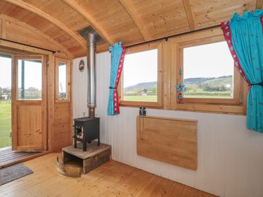 A wooden room with a black wood stove a foldable table blue curtains and windows showing a green landscape at Harris Hut in Bishops Lydeard