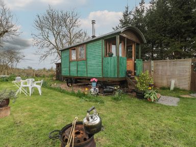 A green wooden cabin on wheels in a garden with lawn chairs a table plants and a kettle on a fire pit at Harri's Hut in Bishop's Lydeard