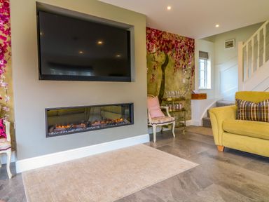 A living room with a wall-mounted television and a fireplace a chair with a pink cushion a sofa and floral wallpaper at Channel View House in Wadebridge
