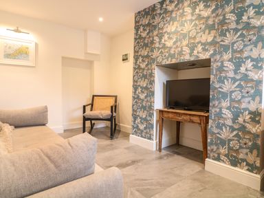 A living room with a gray sofa a wooden chair a TV on a wooden table and floral patterned wallpaper at Channel View House in Wadebridge