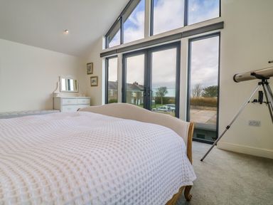 A bedroom with a bed covered with a white blanket a white dresser with a mirror two framed pictures a telescope and large windows at Channel View House in Wadebridge