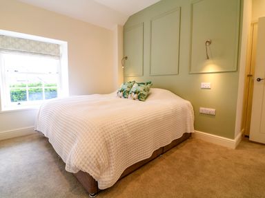 A bedroom with a double bed with white bedding two floral pillows a green accent wall with two reading lights and a window with a blind at Channel View House in Wadebridge