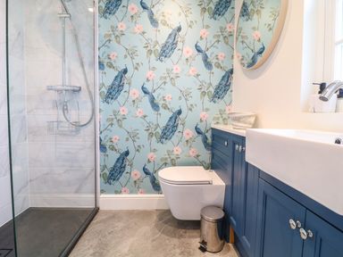A bathroom with a walk-in shower toilet blue cabinets and a floral peacock wallpaper at Channel View House in Wadebridge