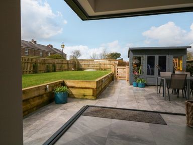 A backyard with a grass lawn bordered by wooden retaining walls a stone patio with a table and chairs and a gray garden shed at Channel View House in Wadebridge