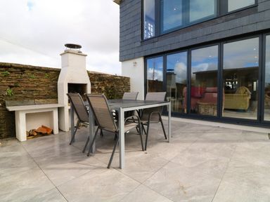 An outdoor patio area with a stone barbecue grill a metal table and six chairs in front of a glass sliding door at Channel View House in Wadebridge
