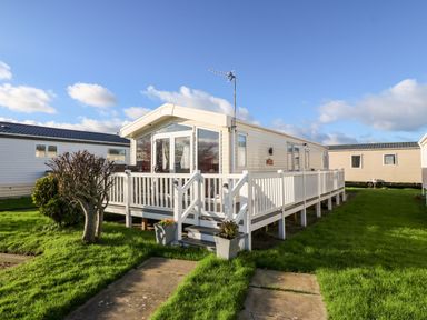 A white mobile home with a wooden porch and railing on a grassy lawn at Snape Lodge in Towyn