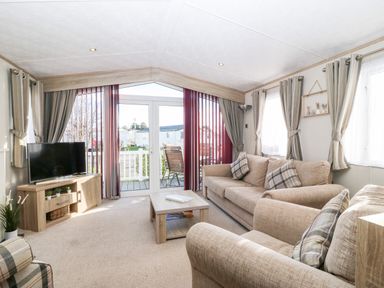 A living room with beige sofas TV wooden table and glass doors leading outside at Snape Lodge in Towyn