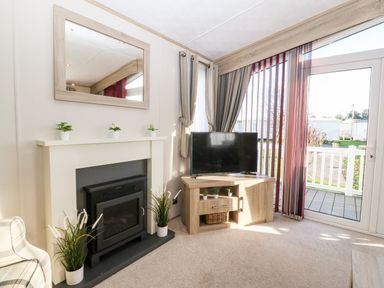 A living room with a fireplace a television on a wooden cabinet and a glass door leading outside at Snape Lodge in Towyn