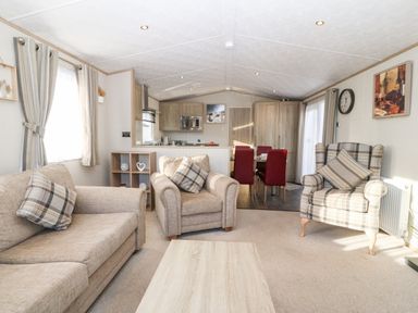 A living room with beige sofas and armchair a wooden coffee table and dining table with red chairs at Snape Lodge in Towyn