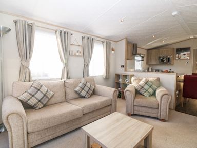 A living room with two beige sofas a wooden coffee table and a view of the kitchen area at Snape Lodge in Towyn