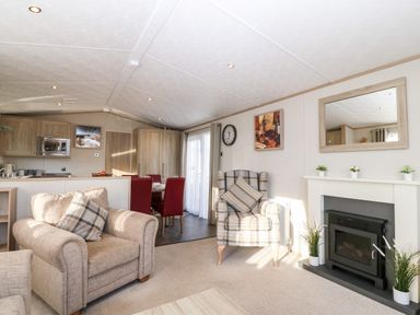 A living room with beige armchairs a dining table with red chairs a fireplace and kitchen cabinets at Snape Lodge in Towyn