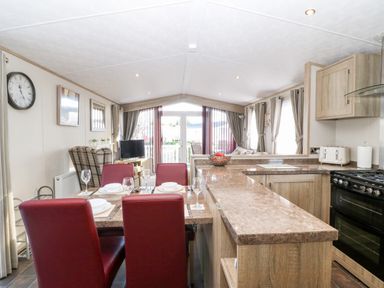 A kitchen and dining area with a table set for four red chairs and a living room with a TV and armchair at Snape Lodge in Towyn