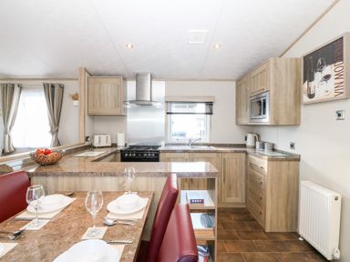 A kitchen with wooden cabinets a stove and a dining table with red chairs at Snape Lodge in Towyn