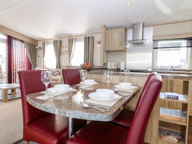 A dining table set with bowls, plates, cutlery, and wine glasses with red chairs in a kitchen and living area at Snape Lodge in Towyn