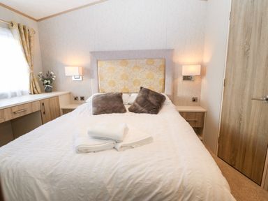 A bedroom with a bed two pillows towels bedside tables wall lights a window with curtains and a wooden door at Snape Lodge in Towyn