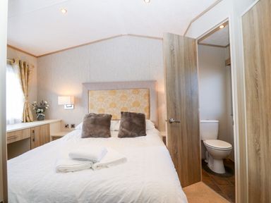 A bedroom with a bed two pillows folded towels bedside tables a window with curtains a vase with flowers and an open door showing a toilet at Snape Lodge Towyn