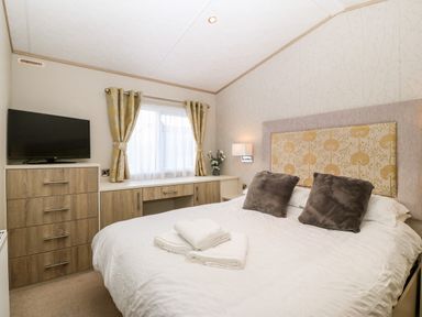 A bedroom with a double bed, two dark pillows, folded towels on the bed, a television on a chest of drawers, and a window with curtains at Snape Lodge in Towyn
