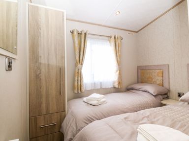 A bedroom with two single beds between a wooden wardrobe and a window with curtains at Snape Lodge in Towyn