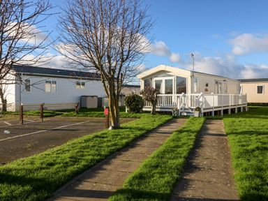 A driveway leading to a beige mobile home with a white railing and trees surrounding it at Snape Lodge in Towyn