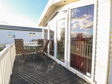 An outdoor deck with a table and four chairs beside a building with large glass doors and windows at Snape Lodge in Towyn