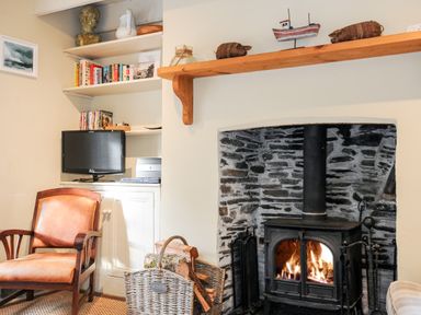 A living room corner with a wood-burning stove in a stone fireplace a wooden chair a TV on shelves and a basket of firewood at Lowen Cottage in Boscastle