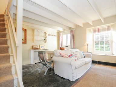 A living room with a white sofa and pillows near a window with curtains and a staircase on the left at Lowen Cottage in Boscastle