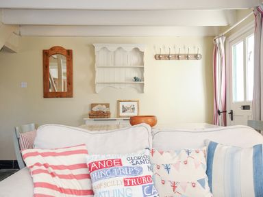 A living room with a white sofa with colorful cushions a wooden mirror a white wall shelf a coat rack and a door with curtains at Lowen Cottage in Boscastle
