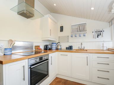 A kitchen with white cabinets wooden countertops a built in oven a stovetop a sink dish rack with plates and mugs and a magnetic knife holder at Lowen Cottage in Boscastle