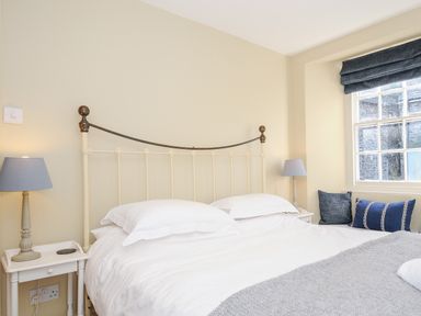 A bedroom with a metal bed frame white bedding bedside tables with lamps and a window with cushions at Lowen Cottage in Boscastle