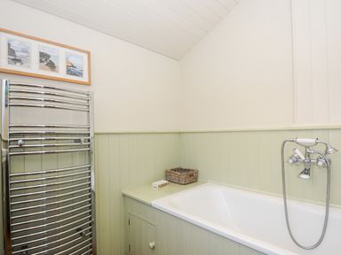 A bathroom with a white bathtub green paneling a chrome towel radiator and framed pictures above at Lowen Cottage in Boscastle