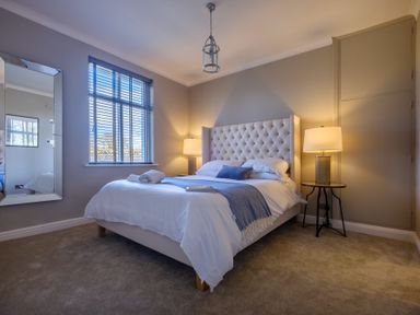 A bedroom with a bed with a tufted headboard two bedside lamps a window with blinds and a wall mirror at Gate House in Killington near Sedbergh
