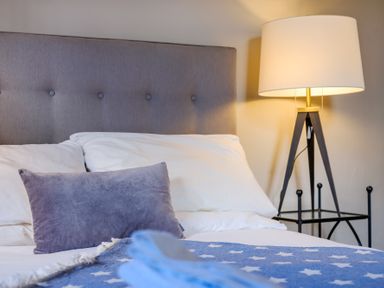 A bed with a gray headboard white pillows a purple cushion a blue blanket with white stars and a lit lamp on a black stand