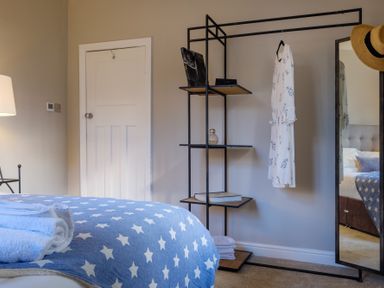 A bedroom corner with a bed covered with a star-patterned blanket and towels a shelving unit with a hanging white dress and a full-length mirror with a hat at Gate House in Killington near Sedbergh