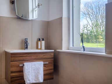 A bathroom sink with a wooden cabinet a white towel two pump bottles under a mirror and a window with a view of trees and grass at Gate House in Killington near Sedbergh