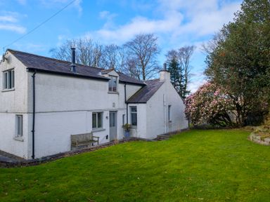 A white two-story house with a slate roof and a wooden bench outside in a garden with green grass and flowering trees at Gate House in Killington near Sedbergh