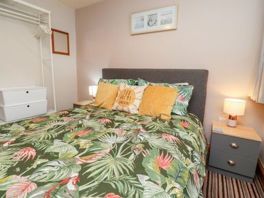A bedroom with a bed with tropical leaf bedding two yellow pillows and one white pillow with text there are nightstands with lamps and a clothing rack with hangers at The Nook in North Sunderland near Seahouses