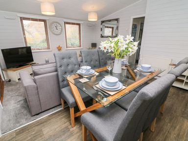 A dining area with a glass table, grey chairs, and a vase of white flowers next to a living room with a sofa and TV at Tree View Lodge in Troutbeck Bridge