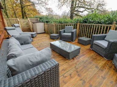 An outdoor wooden deck with grey rattan furniture including armchairs a sofa a coffee table and loungers at Tree View Lodge in Troutbeck Bridge