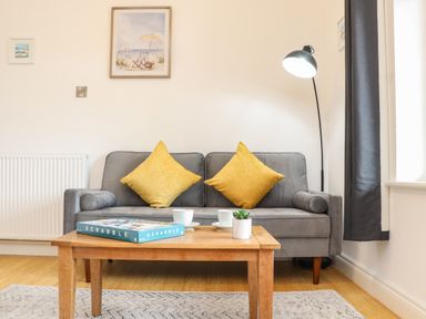 A living room with a gray sofa with yellow cushions a wooden coffee table with a Scrabble game two cups and a small plant a tall floor lamp and framed pictures on the wall at Lakeside in Falmouth