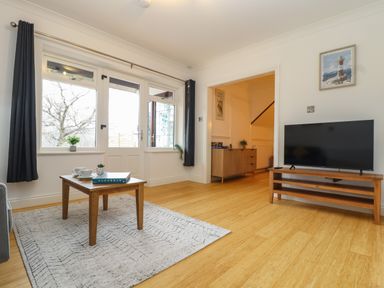 A living room with a wooden coffee table on a rug a television on a wooden stand and large windows with curtains at Lakeside in Falmouth